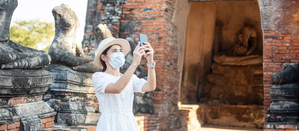 Mid adult woman wearing mask photographing while standing against ancient structure