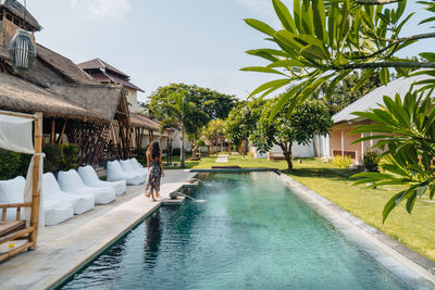 Back view of anonymous female tourist strolling between pool and sunbeds during summer trip in sunlight