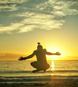 Rear view of man levitating at beach against sky during sunset