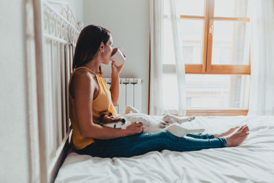 Young woman with dog drinking coffee in cup while sitting on bed