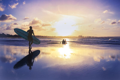 Full length of man walking with surfboard by silhouette people sitting at beach