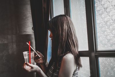 Woman holding drink and using phone while standing by window at home