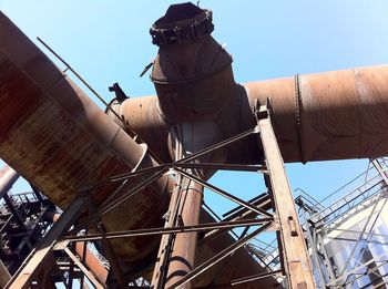 Low angle view of large elevated rusty pipes in decommissioned steel mill against clear blue sky