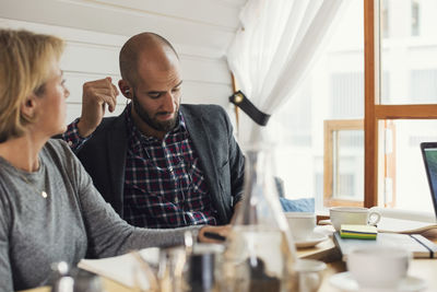 Mid adult businessman wearing in-ear headphones while sitting by female colleague in office