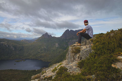 Side view of man sitting on mountain against cloudy sky