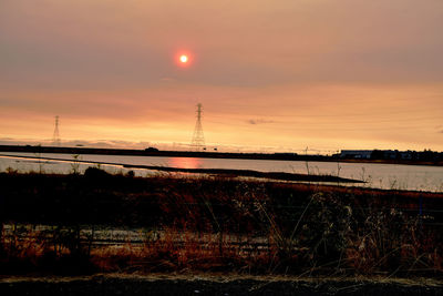 Silhouette electricity pylon by river against sky during sunset
