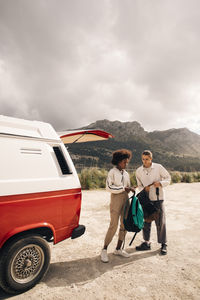 Male and female friends holding backpacks while standing near van against mountains