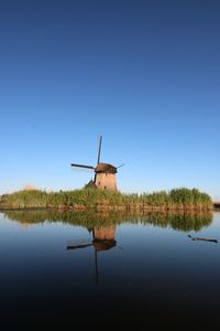 Traditional windmill by lake against clear blue sky