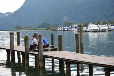 People sitting on pier over lake against mountains