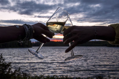 Cropped image of couple toasting wineglass by river during sunset