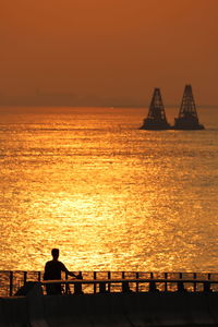 Silhouette people sitting on sea against orange sky