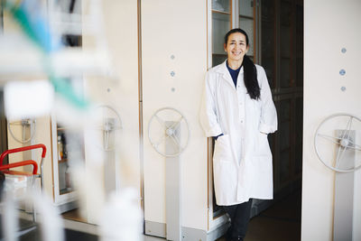 Portrait of confident mature female teacher standing with hands in pockets against storage cabinets in university