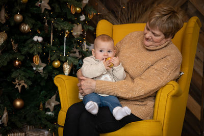 Smiling grandmother sitting with granddaughter by christmas tree at home