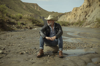 Adult man in cowboy hat sitting on rock along river flows in desert. almeria, spain