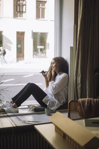 Smiling female design professional talking on speaker phone while sitting on window sill at office