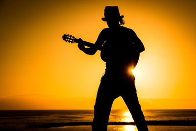 Silhouette man playing guitar at beach against orange sky