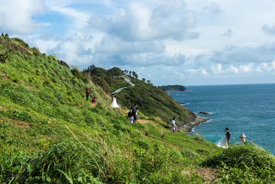 Photographers with bride and groom on grassy hill by sea against sky