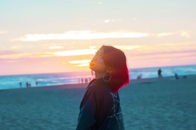 Portrait of young woman with dyed hair standing at beach against sky during sunset