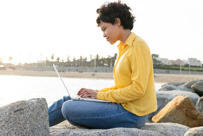 Side view of young female in casual clothes sitting on seafront rock and browsing laptop while spending time on city seashore