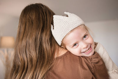 Girl putting head on mother shoulder smiles and looks away camera.