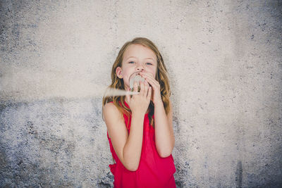 Cute girl playing with tin can phone while standing against wall