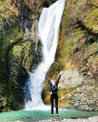 Full length of woman with arms raised standing against waterfall
