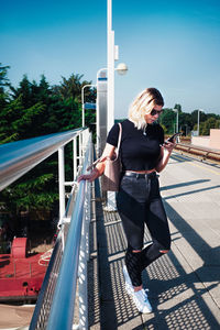 Woman using phone while standing by railing against sky