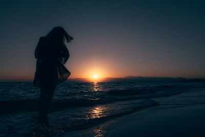 Rear view of woman standing at beach against sky during sunset