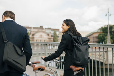 Smiling businesswoman looking at businessman while walking with bicycles on bridge in city