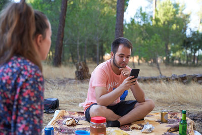 Man using mobile phone while sitting at forest with woman