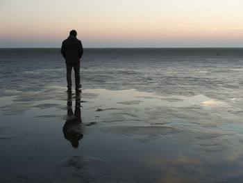 Rear view of man standing in sea against sky during sunset