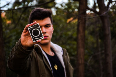 Portrait of young man holding cards while standing against trees