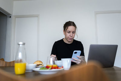 Young woman using smart phone while sitting with laptop and breakfast at dining table