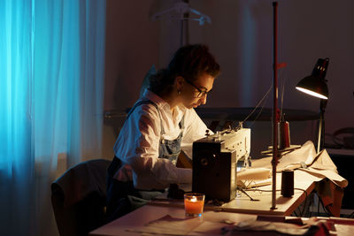 Busy designer woman working late at night in atelier with sewing machine, fabric and paper patterns