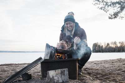 Happy woman warming up boiling a kettle on a camp fire at the beach