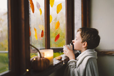 Boy wrapped in a blanket drink hot tea and looks out the open window at the wonderful autumn nature