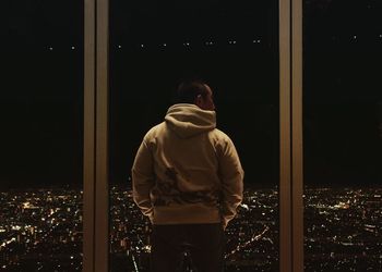 Rear view of man looking at illuminated city from window