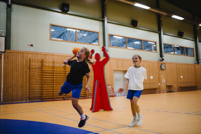 Girl jumping while playing handball with female friend in sports court