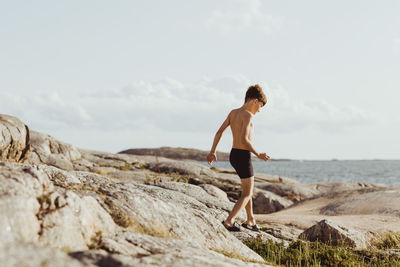 Full length of shirtless man on rock at beach against sky