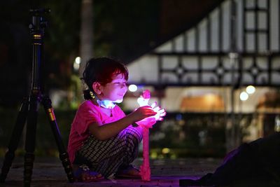 Side view of woman sitting against illuminated lights at night