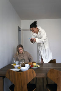 Happy woman photographing breakfast while standing on chair by girlfriend at home