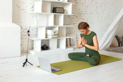 A woman in a green sports jumpsuit, in a bright room, online performs a yoga exercise