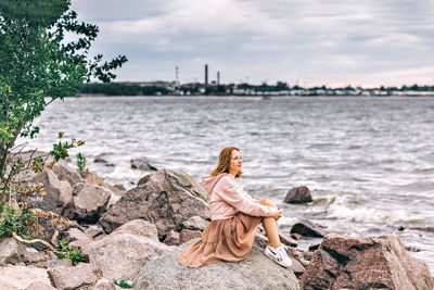 Rear view of woman sitting on rock looking at shore against sky