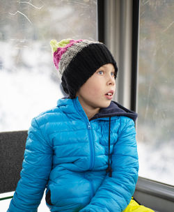 A boy in a blue jacket and a knitted hat sits in a booth by the winter window and dreams. 