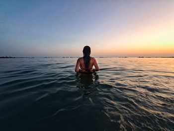 Rear view of girl standing in sea against sky during sunset