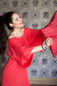 Young woman holding red pillow while standing against wall