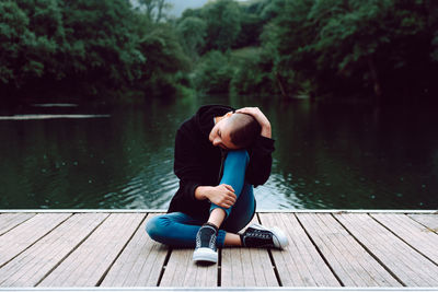 Unique female hipster with short hairstyle in casual clothing sitting with crossed legs on wooden pier of pond with green trees on blurred background