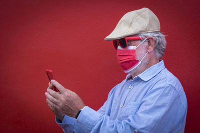 Senior man wearing mask using smart phone against red background