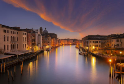 Illuminated buildings by river against sky at sunset