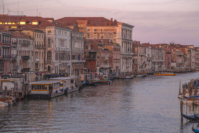 Boats in canal by buildings in city against sky during sunset